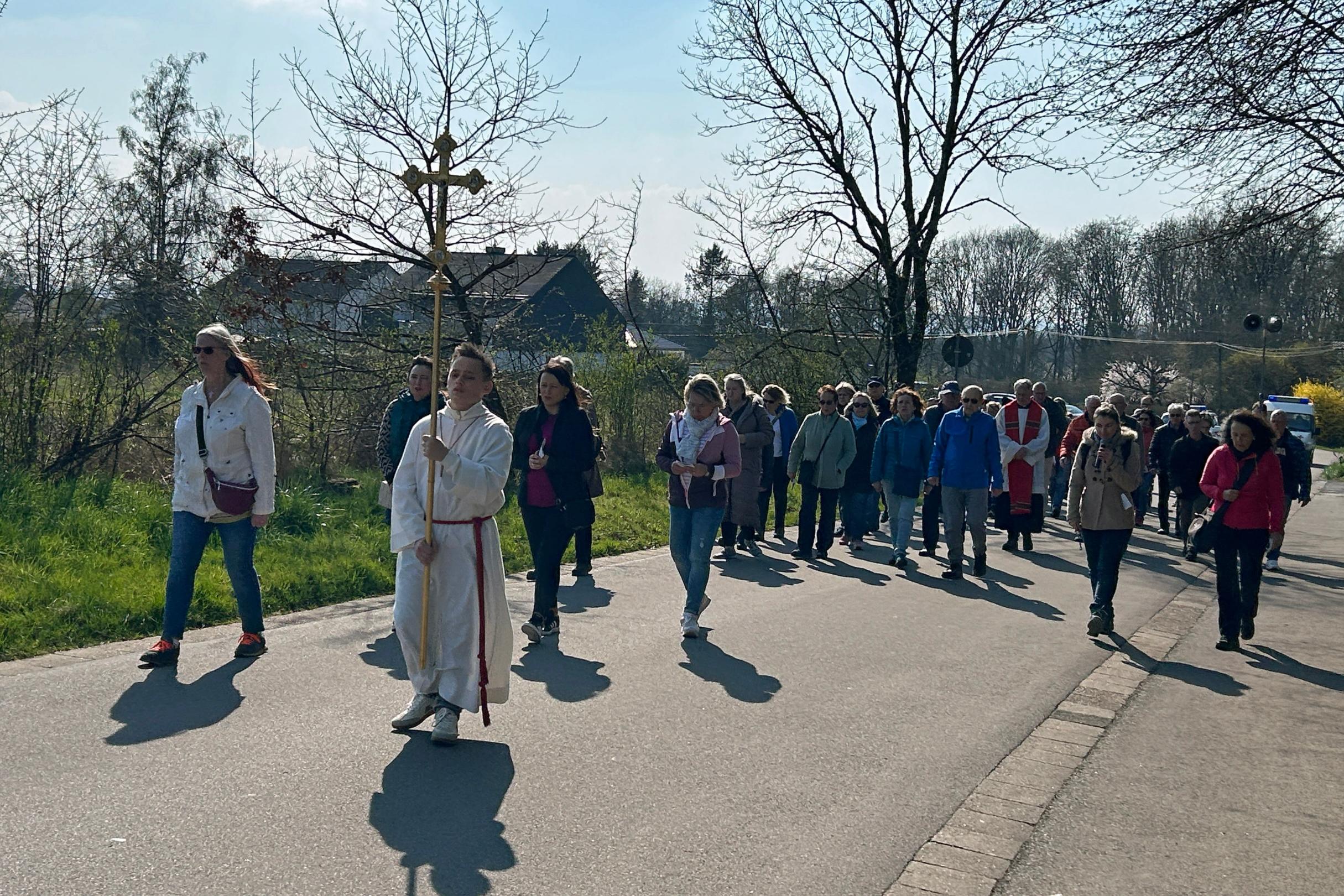 Eine größere Gruppe von Menschen geht bei sonnigem Wetter auf einer asphaltierten Straße zwischen Wiesen und Bäumen. An der Spitze der Prozession trägt eine Person ein großes Vortragekreuz. Hinter dieser Person folgt eine dicht geschlossene Gruppe von Teilnehmenden, einige in Jacken, andere mit Taschen oder Rucksäcken. Rechts und links des Weges stehen noch kahle Bäume und frühe Frühlingssträucher. Im Hintergrund sind einige Häuser und ein freies Feld zu sehen. Die Szene wirkt wie eine gemeinschaftliche religiöse Prozession oder ein gemeinsamer Weg durch die ländliche Umgebung.
