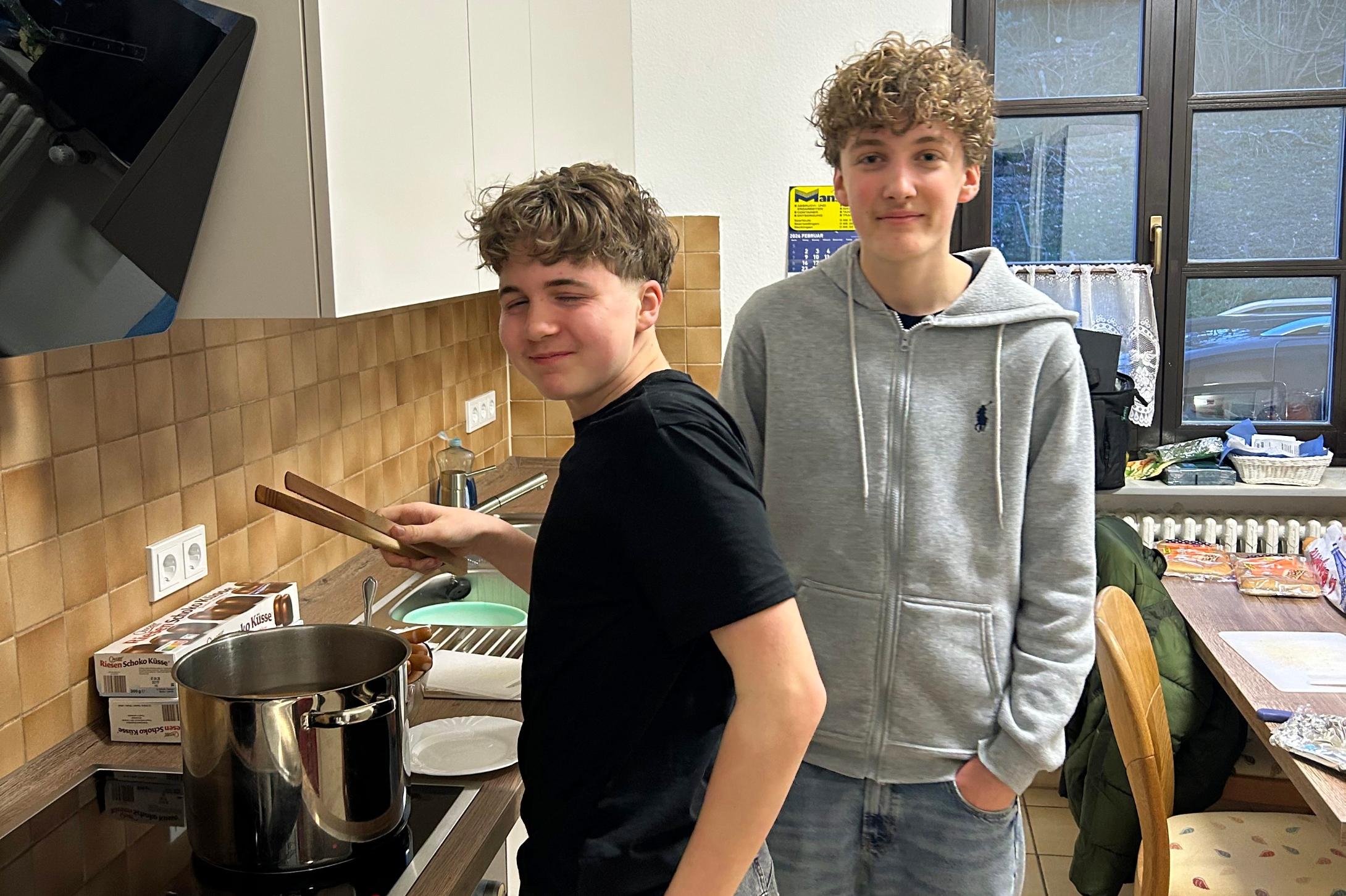 Zwei Jugendliche stehen in einer Küche. Eine Person steht am Herd und rührt mit Küchenzangen in einem großen Edelstahlkochtopf auf der Arbeitsplatte. Daneben liegen ein Topfdeckel und Zutaten wie Hotdog-Brötchen. Die zweite Person steht leicht seitlich dahinter, mit den Händen in den Hosentaschen. Im Hintergrund sind Küchenschränke, ein Fenster, ein Esstisch mit Stühlen und weitere Lebensmittel zu sehen.